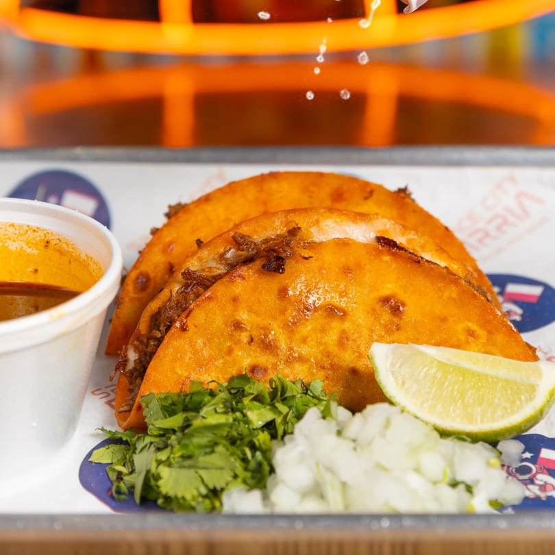 Three Space City Birria tacos with a side of consommé, cilantro, diced onions, and a lime wedge.