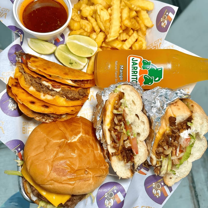 A Space City Birria meal with birria tacos, burger, tortas, fries, consommé, limes, and a Jarritos.