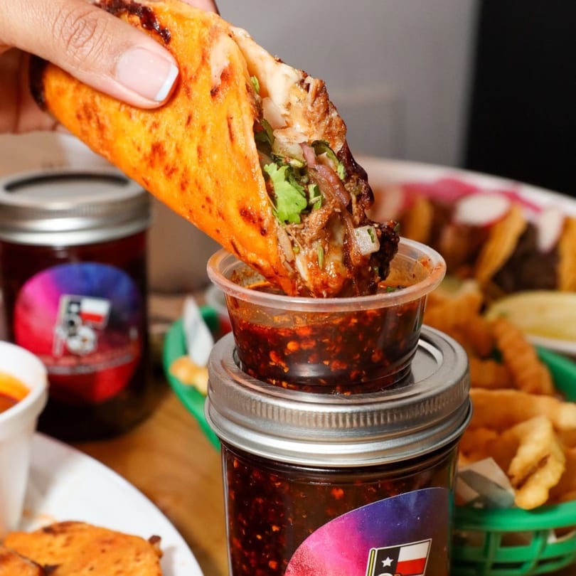 A hand dips a Space City Birria taco, filled with meat and cilantro, into a small cup of birria consommé.