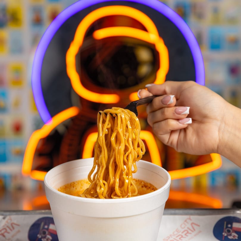 Hand holding a fork lifting birria ramen noodles from a white cup, with a neon astronaut in the background.